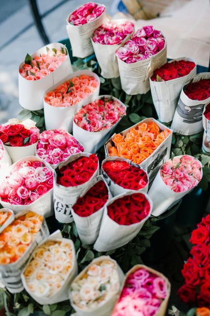Various floral bouquet arrangements displayed on a table, featuring fresh roses in shades of pink, peach, red, and cream, wrapped in white and printed paper with floral motifs. The bouquets are tightl