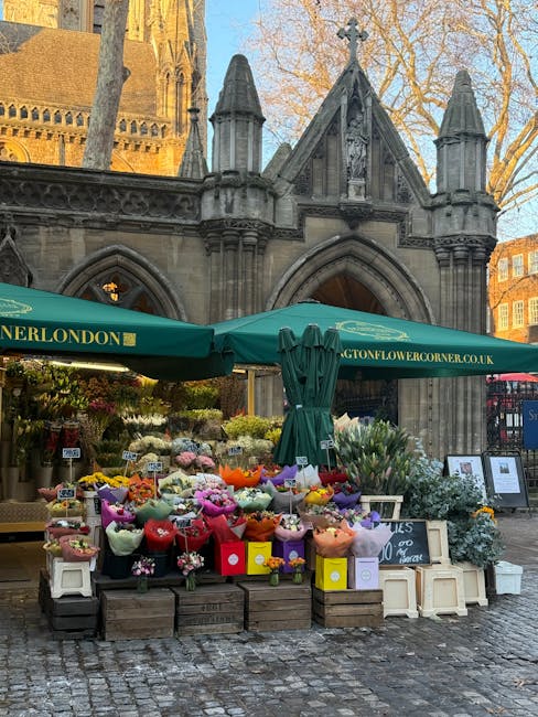 A vibrant outdoor flower stall situated in front of a historic stone building with Gothic architectural details at Marylebone Station. The stall, covered by an emerald green canopy labeled 'London Flo
