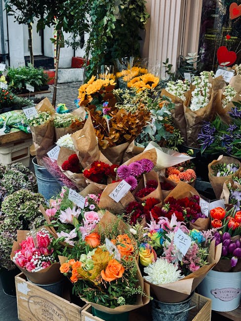 A colorful display of fresh flowers arranged in bouquets and potted plants outside a florist shop. The bouquets include vibrant pink lilies, orange roses, purple irises, white chrysanthemums, and red