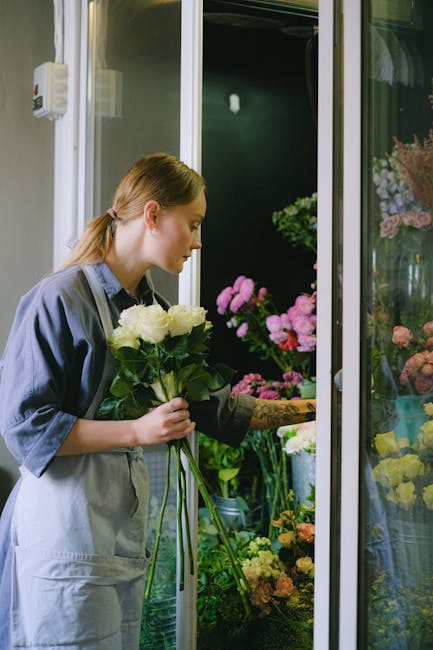 A young woman with light brown hair tied back in a ponytail is holding a bouquet of white roses with green leaves inside a flower shop. She is dressed in a grey apron over a blue shirt and is examinin