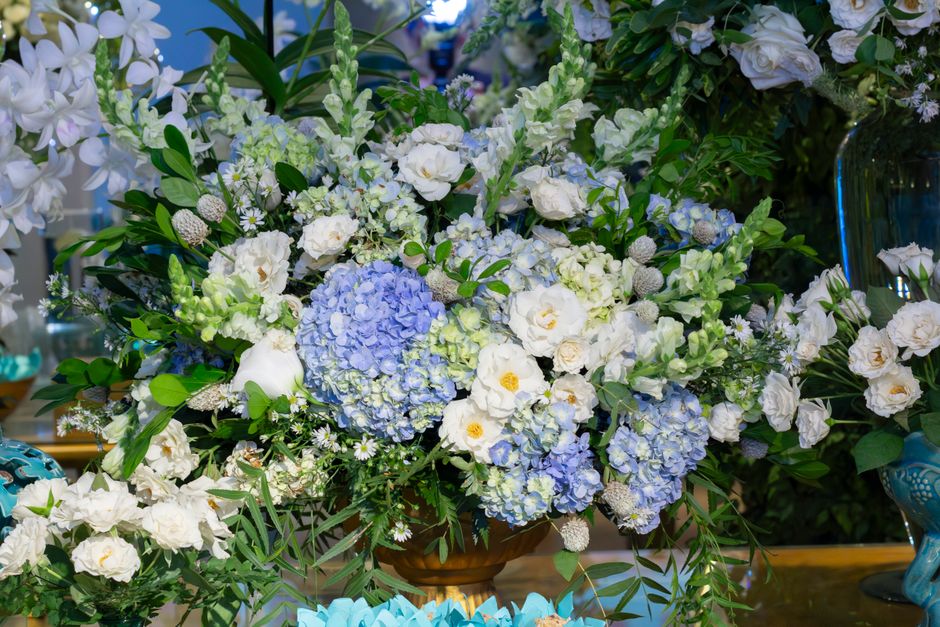 A large floral arrangement featuring a mix of white and pale blue flowers, prominently including clusters of hydrangeas and roses, complemented by various green foliage. The flowers are arranged in a 