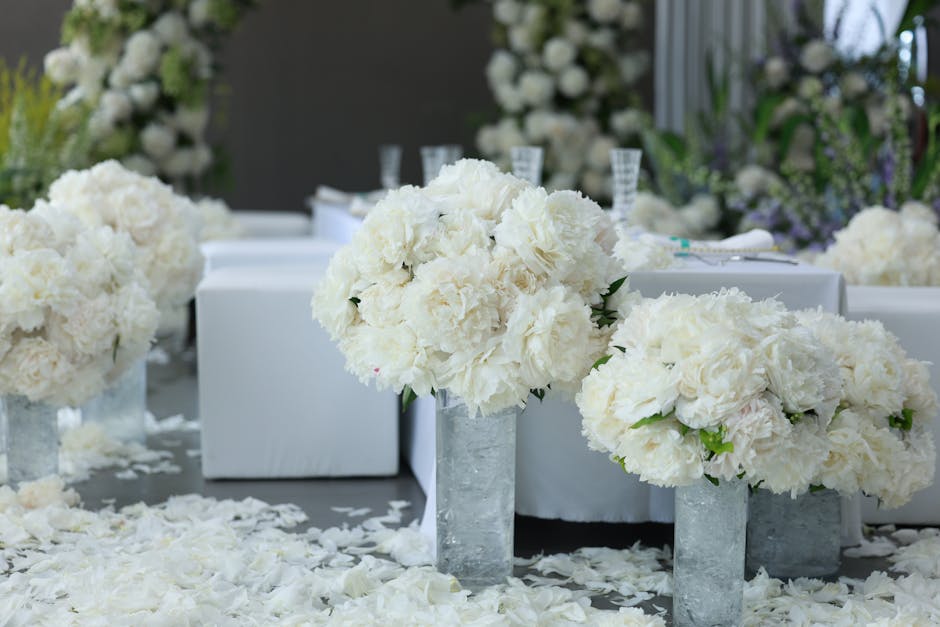 A floral arrangement featuring large, rounded bouquets of white peonies and hydrangeas placed in tall, cylindrical glass vases on a table covered with white flower petals. The bouquets are lush and de