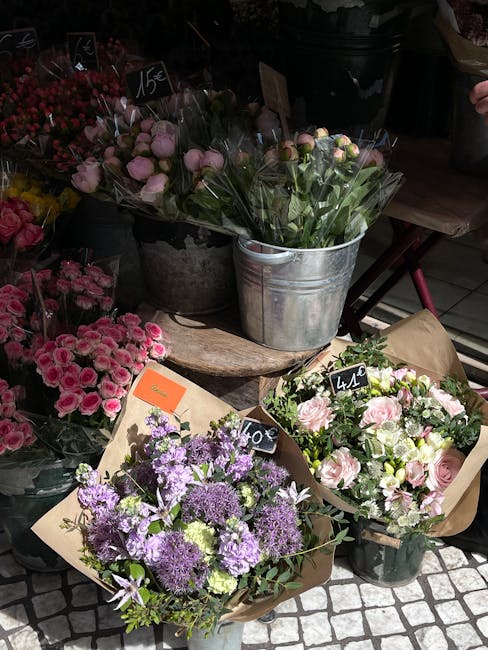 A collection of fresh floral arrangements displayed outdoors at a flower shop, including bouquets of pink roses wrapped in brown paper, clusters of purple and lavender flowers in a gray watering can,