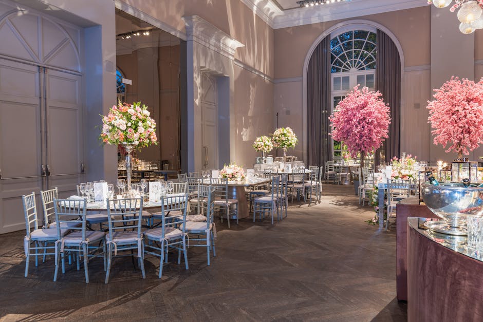 The image depicts a spacious, elegantly decorated room with high ceilings and large arched windows, filled with natural light. Several round tables are arranged throughout the space, each adorned with