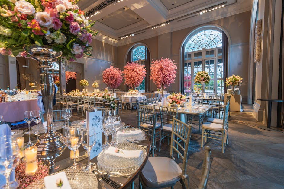 An elegant wedding reception set inside a bright, spacious hall with large arched windows and high ceilings. The tables are decorated with floral arrangements featuring pink cherry blossom trees as fo